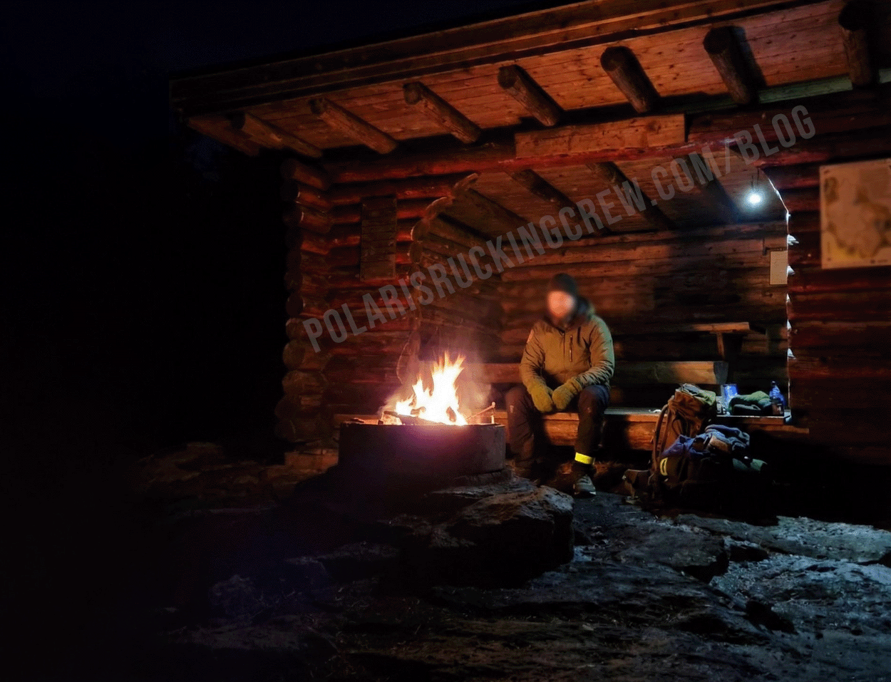 A person sitting alone beside a fire outside a wooden shelter on a winter night